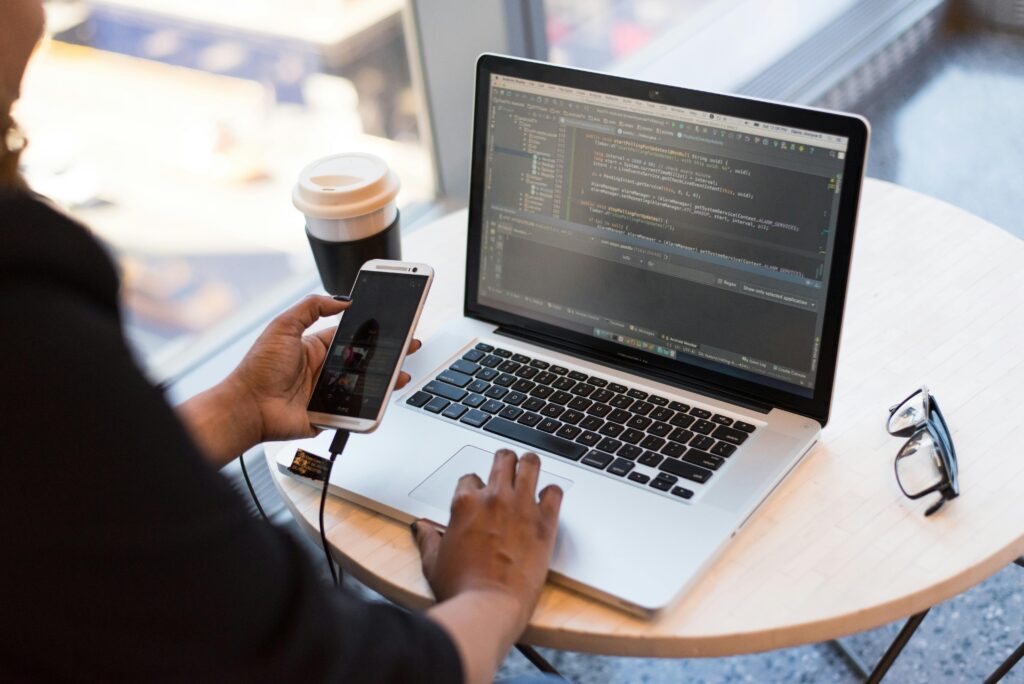 pexels-photo-1181244-1181244 Developer working remotely, coding on a laptop with phone in hand, showcasing modern work culture.